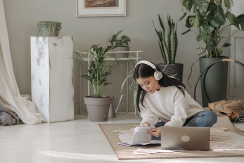 Free Asian woman seated on floor, using laptop and headphones, surrounded by indoor plants. Stock Photo