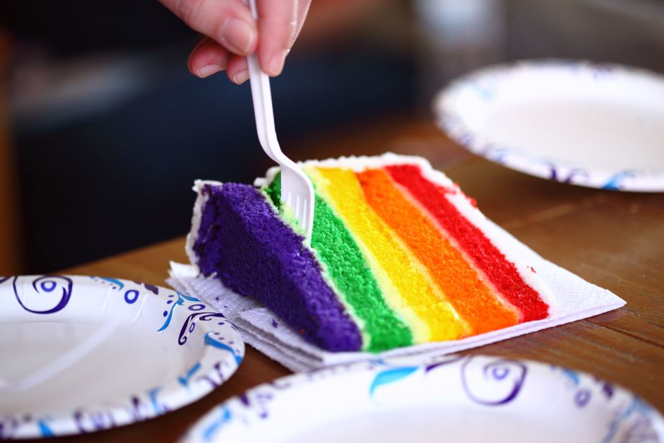 A tall six-layer rainbow cake sliced to show red, orange, yellow, green, blue, and purple layers stacked with white frosting between each, frosted smooth on the outside.