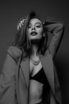 Black and white of young confident female in jacket and bra with necklace looking at camera in light studio