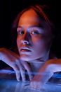 Thoughtful young wet lady looking at camera while leaning on glass container with hands in water in dark room with blue and orange neon lights