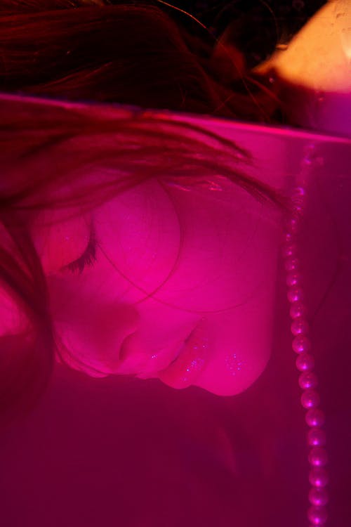 Thoughtful young female with face in glass container with purple liquid and pearl necklace in dark room with closed eyes