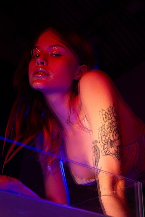 Confident young female in bra with makeup and tattoos on hand near glass container with liquid looking at camera in dark room with neon lights