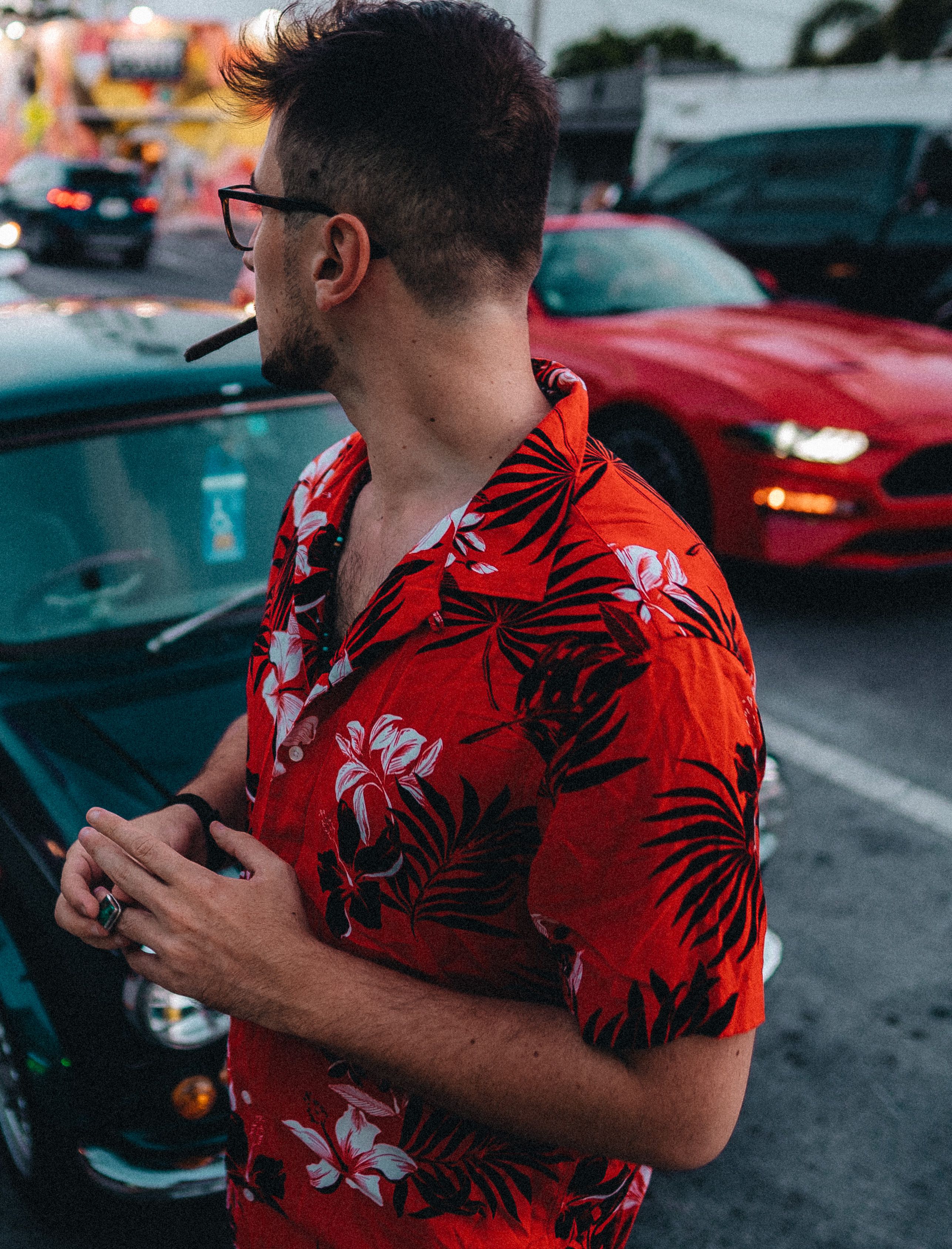 Profile of a man in floral shirt smoking a cigar, standing beside parked cars in Miami.
