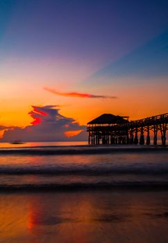Scenic sunset on a tranquil beach with a silhouetted jetty reflecting in calm waters.