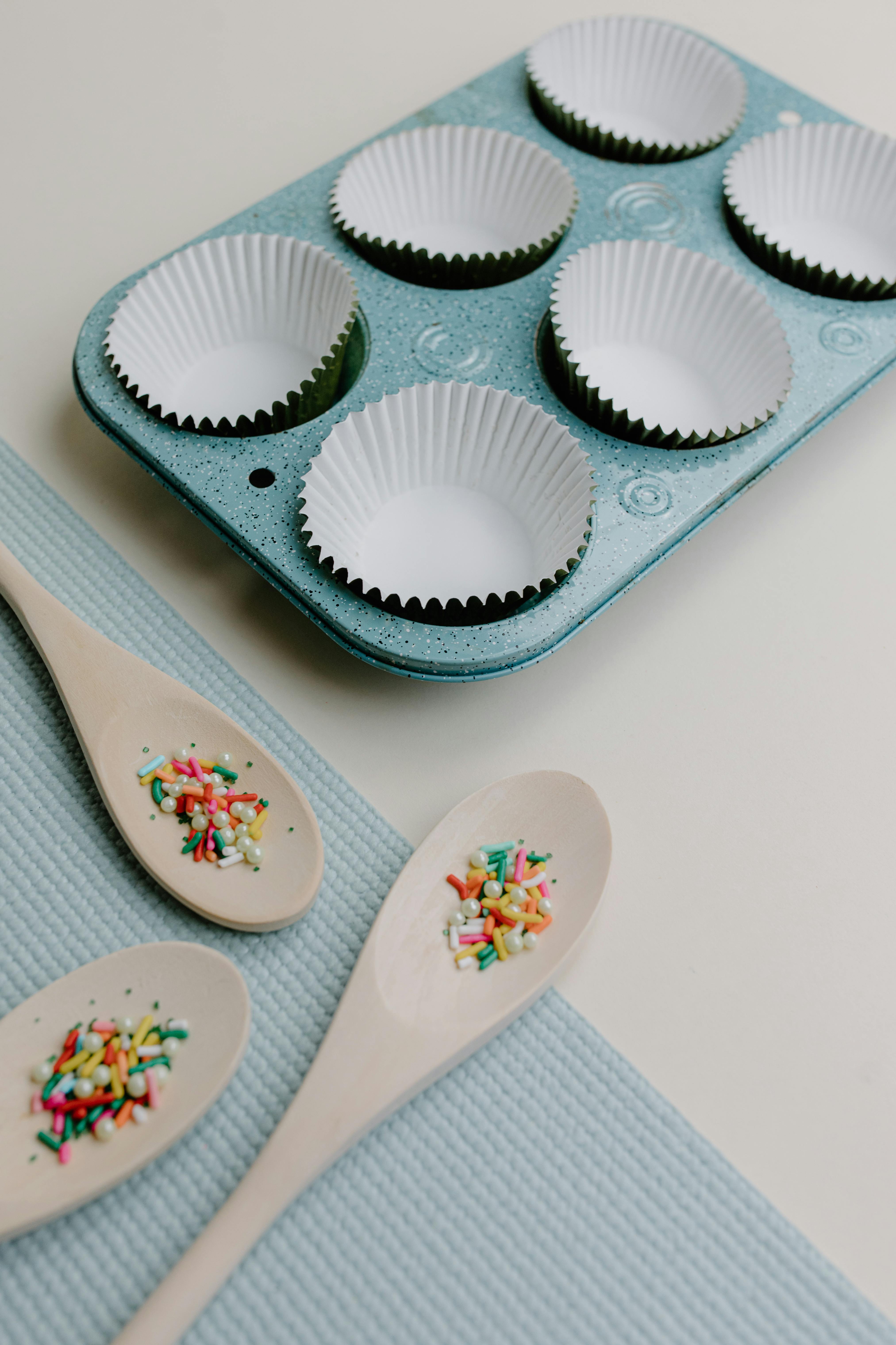 Baking setup with muffin tins, cupcake liners, and sprinkles on wooden spoons.