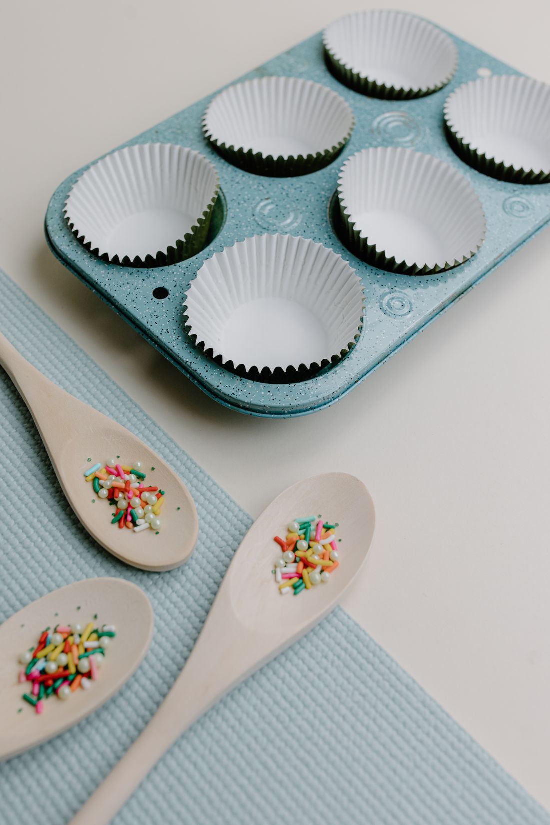 Speckled aqua muffin pan with paper liners and wooden spoons of sprinkles