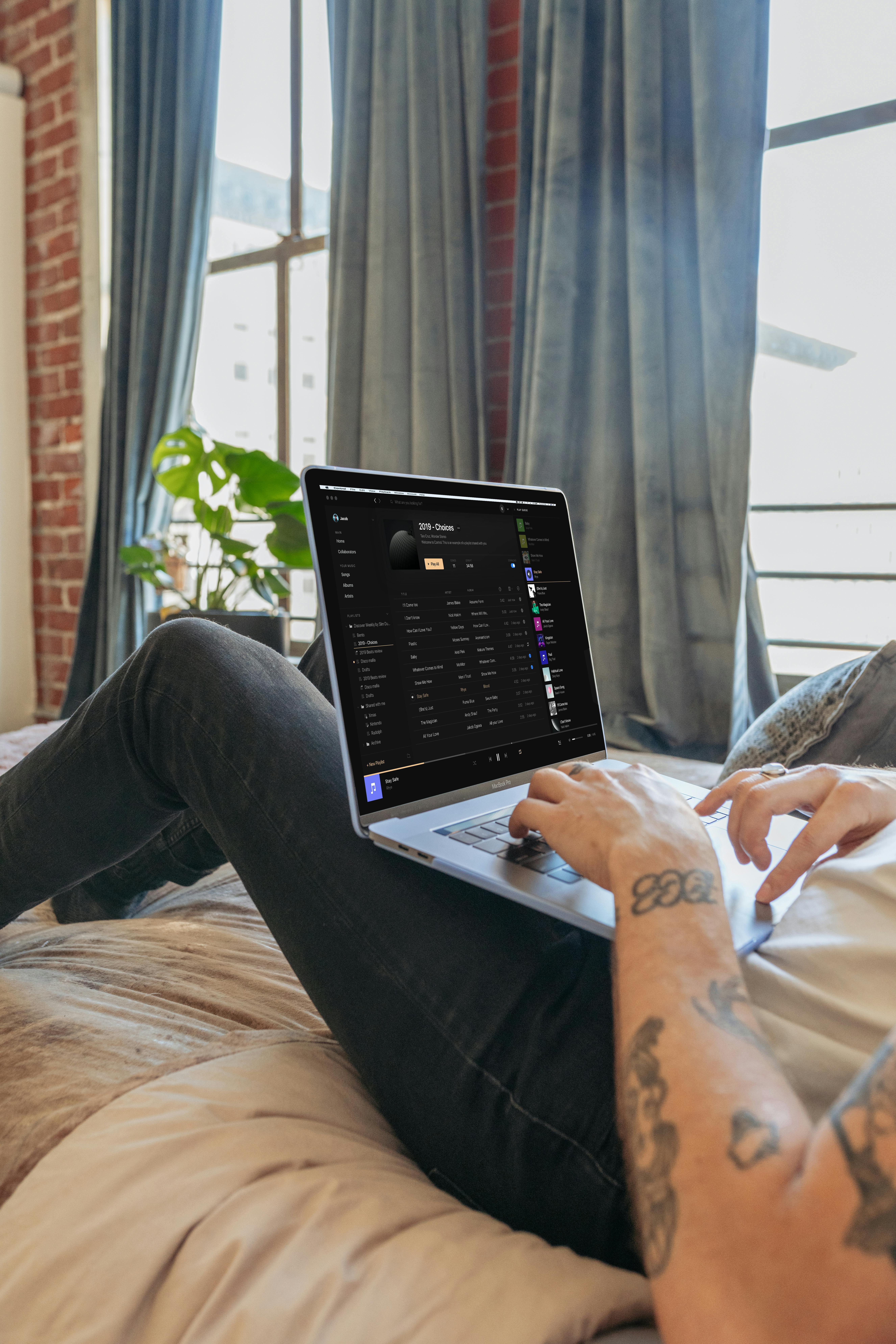 A tattooed person relaxes on a bed browsing a laptop in a cozy bedroom with natural light.