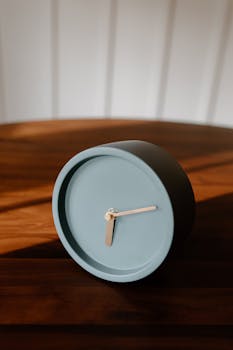 Close-up of a minimalist clock on a wooden table, capturing soft natural light and shadows.