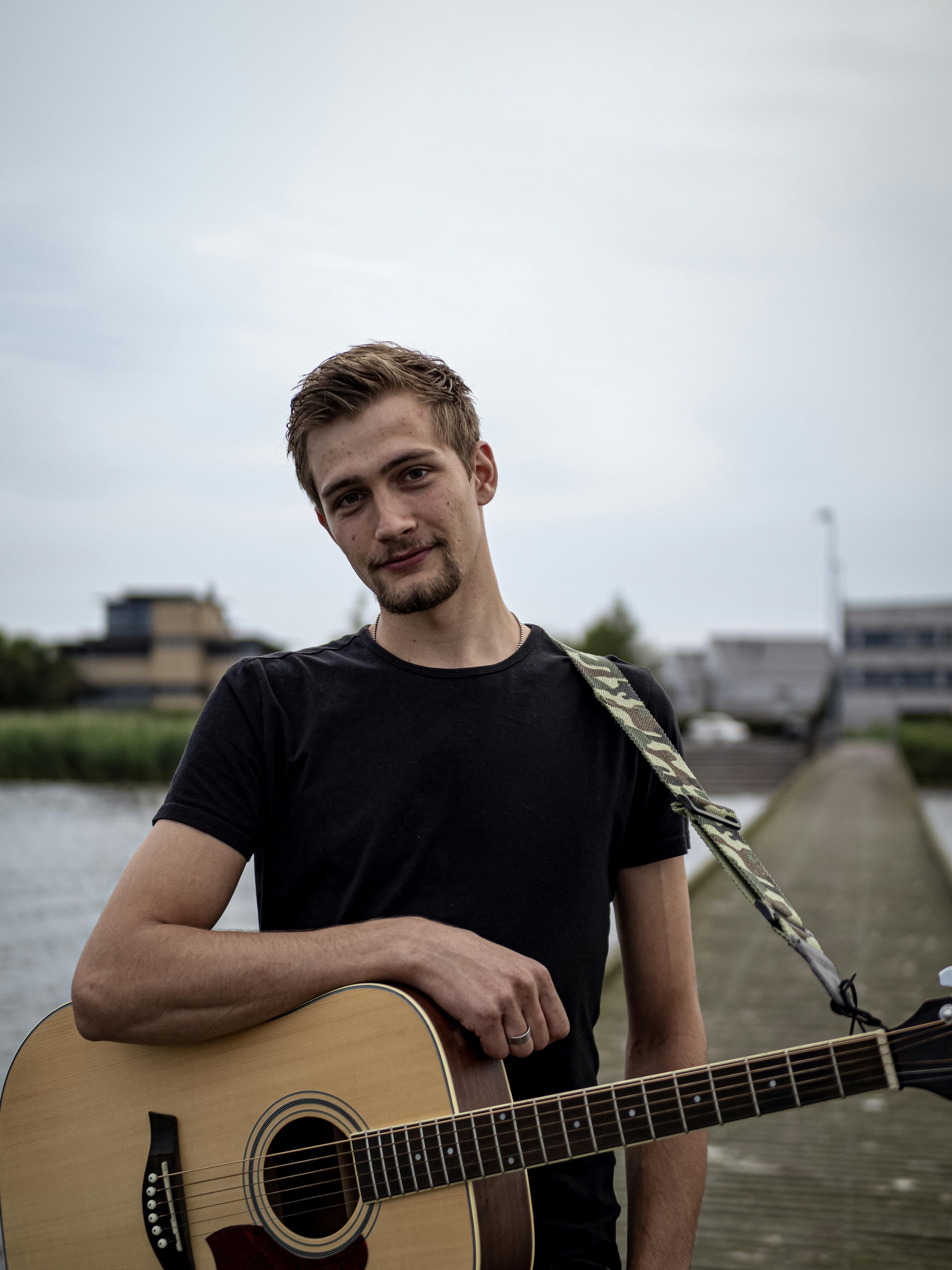 A young man with an acoustic guitar standing on a pier by the sea.