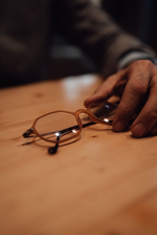 Crop anonymous male touching rims of eyeglasses placed on empty wooden table while sitting in light room on blurred background