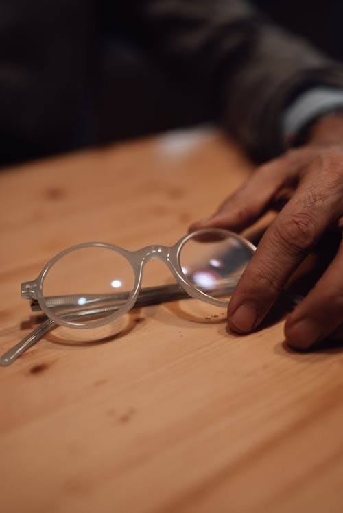 Crop unrecognizable male touching eyeglasses with round shape and gray frame placed on wooden table in room on blurred background