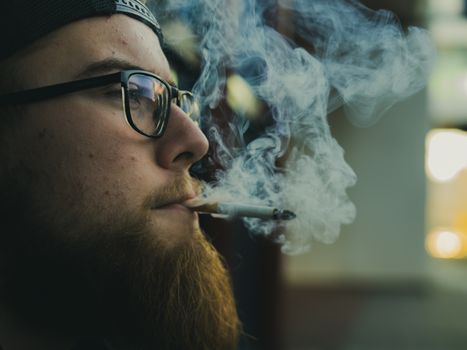 Close-up shot of a bearded man with glasses smoking a cigarette, featuring swirling smoke.