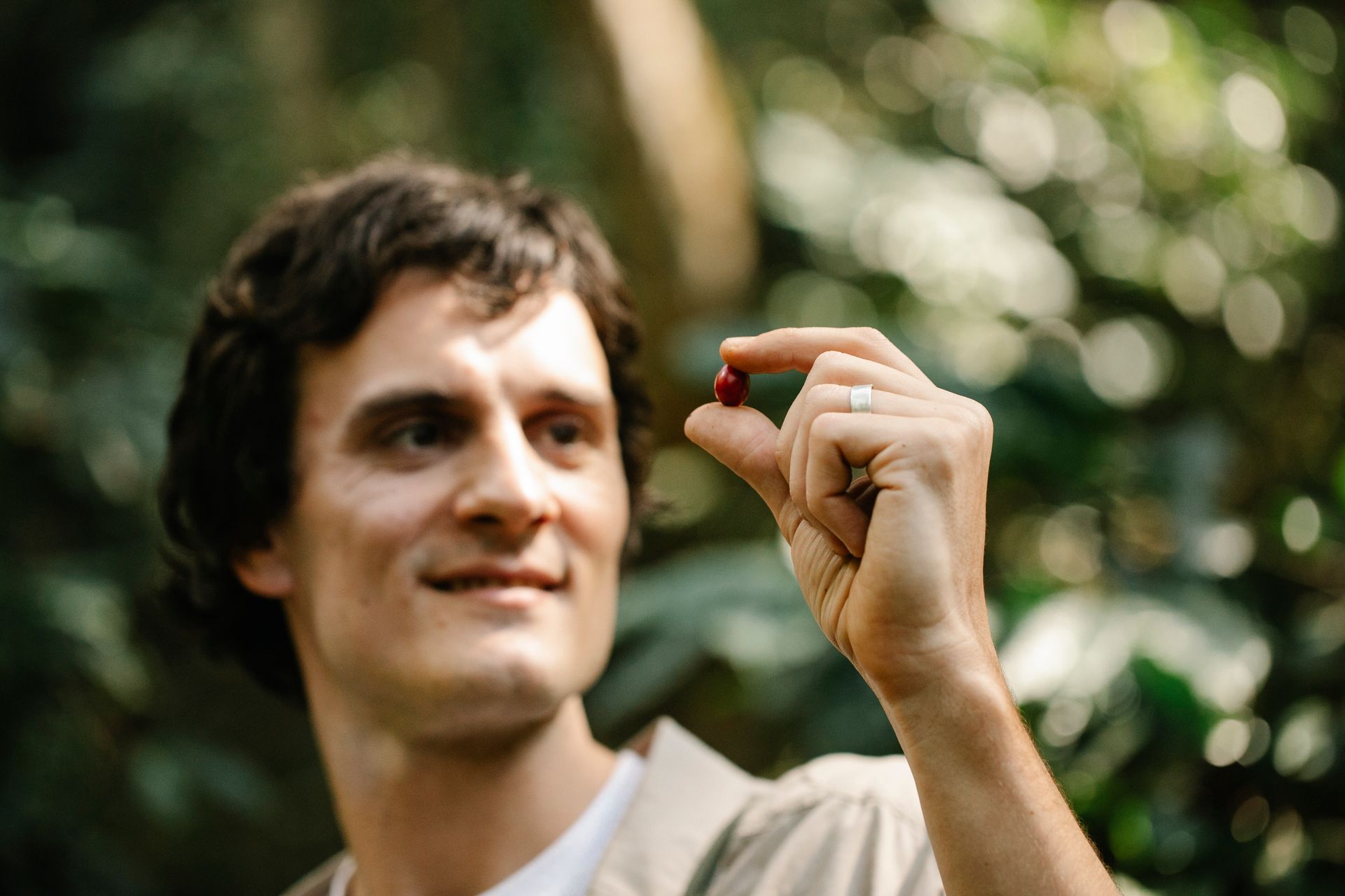 Cheerful adult male planter looking at ripe red coffee cherry in raised hand while standing in sunny abundant garden