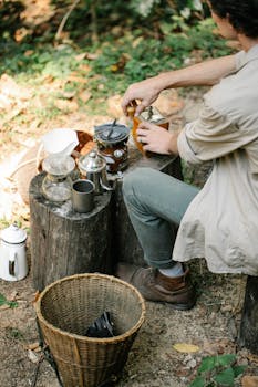 Man prepares coffee outdoors using manual equipment on wooden stumps in rustic setting.