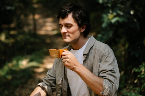 Dreamy male with wooden container enjoying coffee scent on pathway in soft sunlight on blurred background