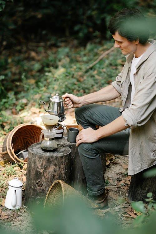 Side view of male harvester with kettle and pour over coffee maker preparing hot beverage on stump