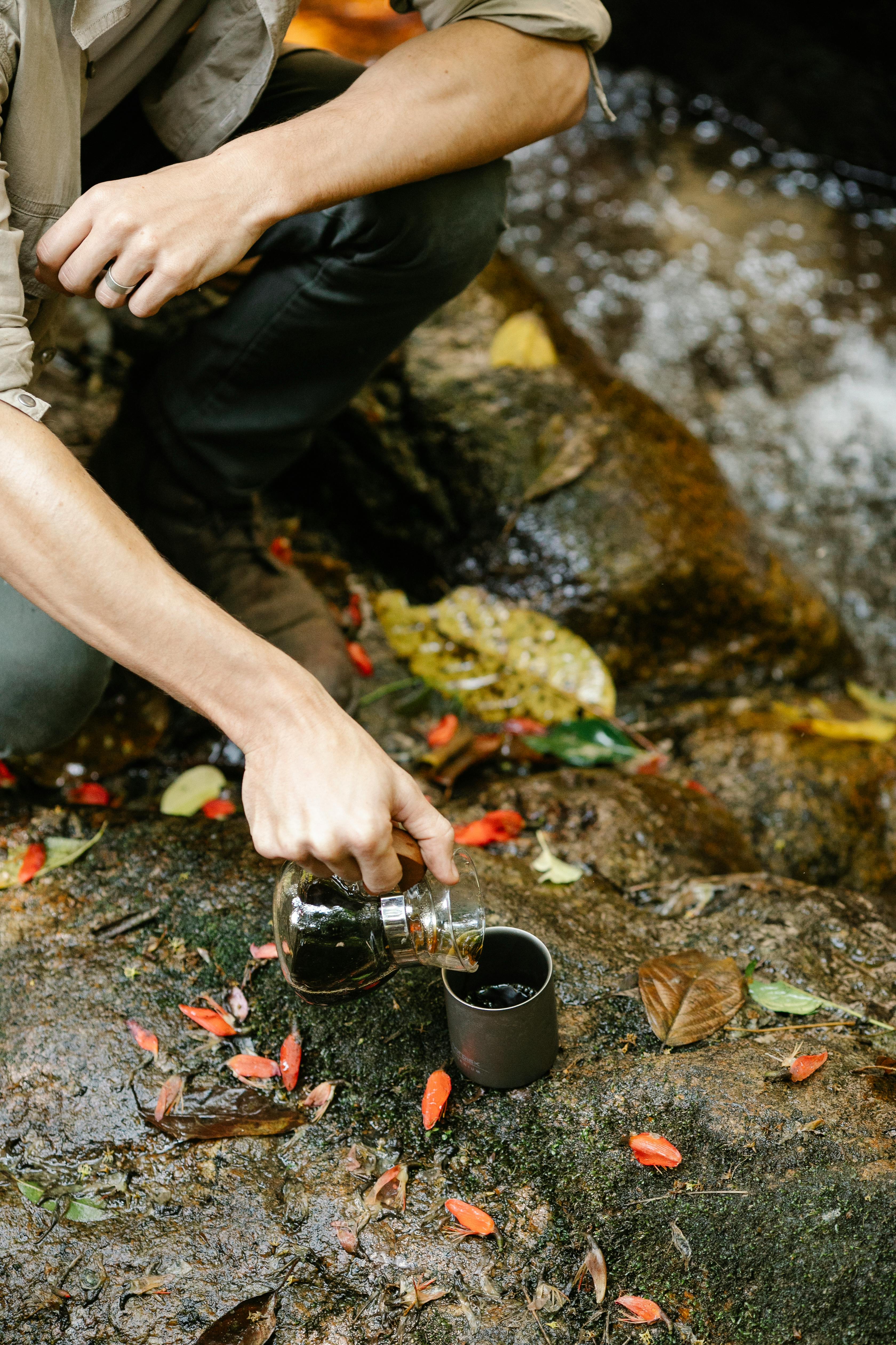 man pouring coffee into metal cup near clean brook
