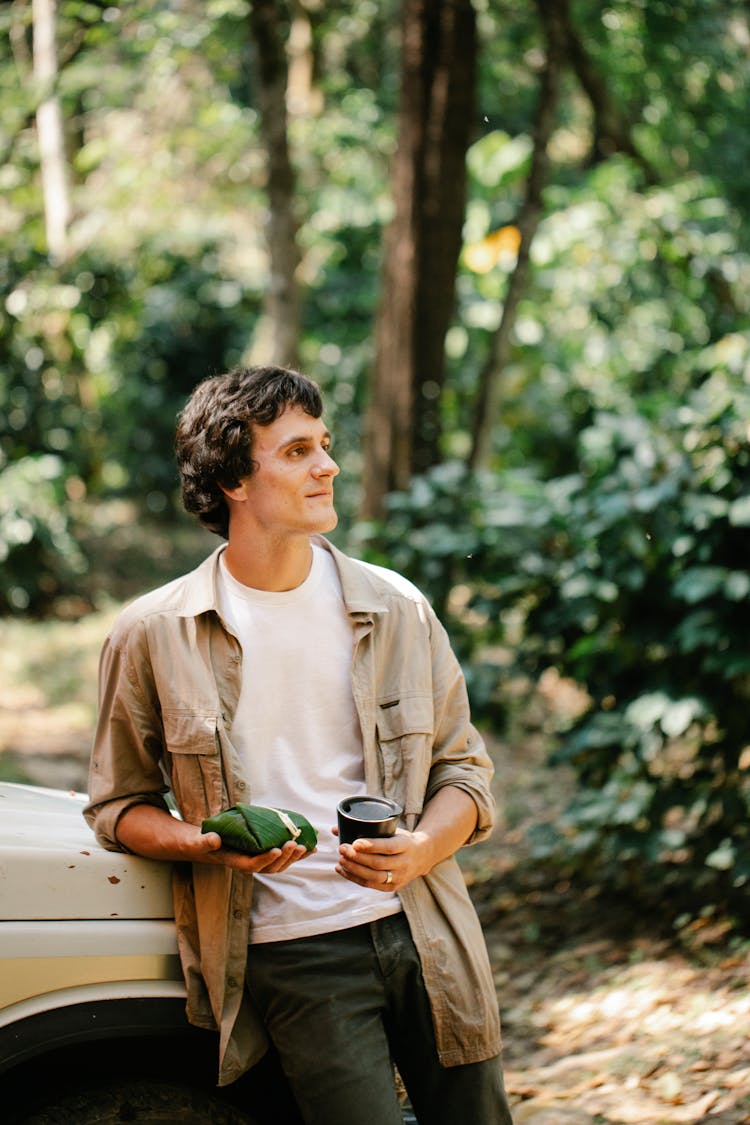 Positive Man With Coffee Leaning On Car In Forest