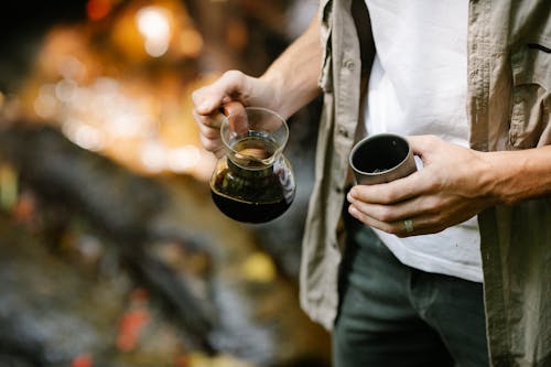 Crop anonymous male camper pouring freshly brewed coffee from chemex coffeemaker into metal mug while resting in autumn forest during trekking
