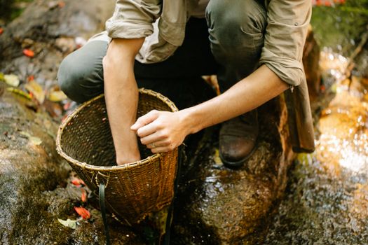 From above of crop anonymous male hiker in casual clothes sitting on haunches and collecting berries into wicker bucket near wild rocky creek in forest