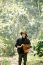 Calm young male harvester in denim jacket and hat holding wicker basket and looking away while standing in lush green vineyard on sunny day