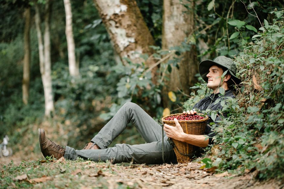 Side view full body of content male in hat with wicker basket full of coffee beans sitting on ground in forest