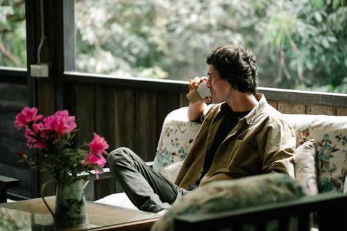 Free Adult male drinking aromatic hot beverage from glass while sitting on couch and looking away against blooming flowers on veranda Stock Photo