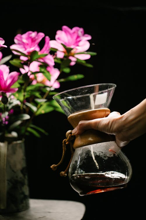 Crop anonymous person with hot drink in glass jug against blossoming flowers on black background