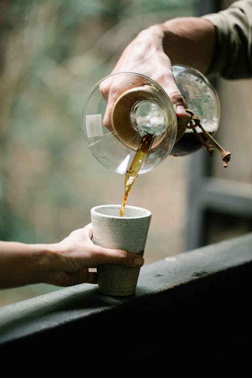 Crop unrecognizable male pouring aromatic hot drink from Chemex coffee maker into glass against female partner on veranda in countryside