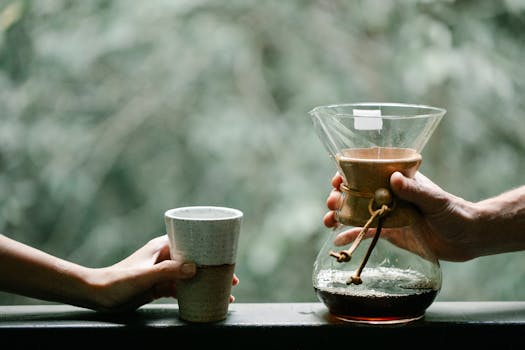 Two hands exchanging freshly brewed coffee in a Chemex outdoors.