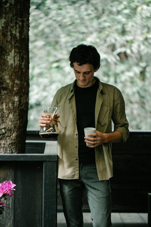 Young male traveler pouring fresh brewed coffee from chemex coffeemaker into cup while standing on wooden terrace of house in lush green forest