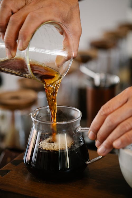 Crop unrecognizable male barista pouring freshly brewed aromatic coffee from glass measuring pitcher into hot at counter