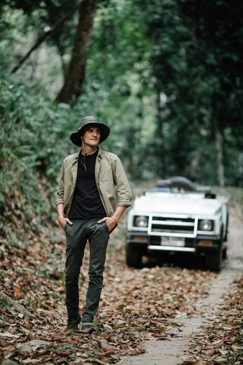Positive young man walking in autumnal forest near car parked on narrow path