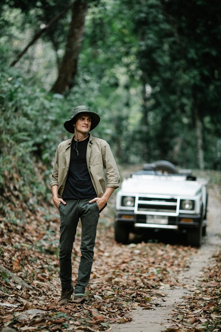 Positive Young Man Walking In Autumnal Forest Near Car Parked On Narrow Path