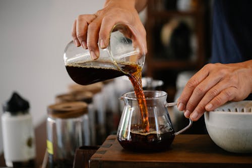 Free Crop faceless male worker pouring aromatic hot coffee from glass measuring pitcher into pot placed on wooden counter in cafe Stock Photo