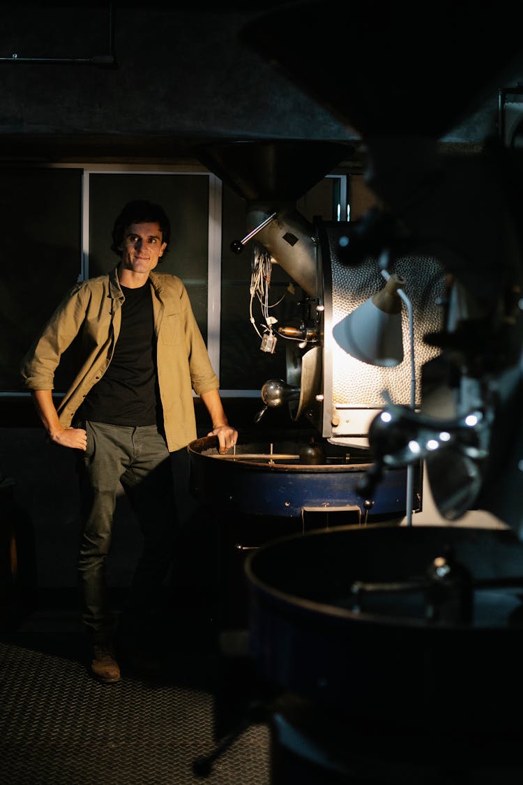 Young Guy Standing In Dark Room With Coffee Roasting Machine In Factory