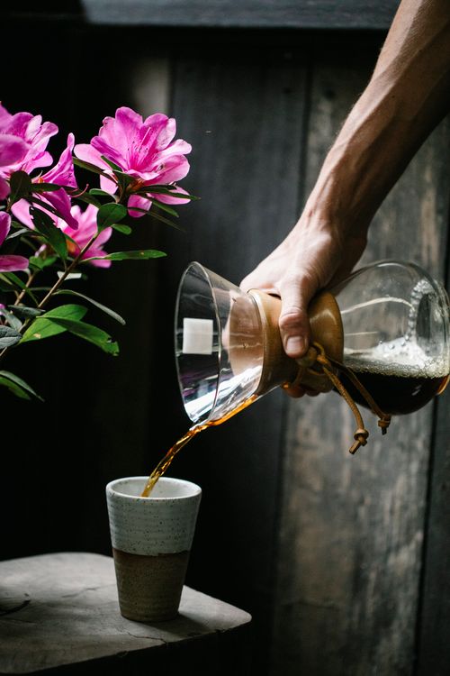 Free Crop unrecognizable guy pouring freshly brewed coffee from chemex glass carafe into cup placed on table near bunch of delicate flowers Stock Photo