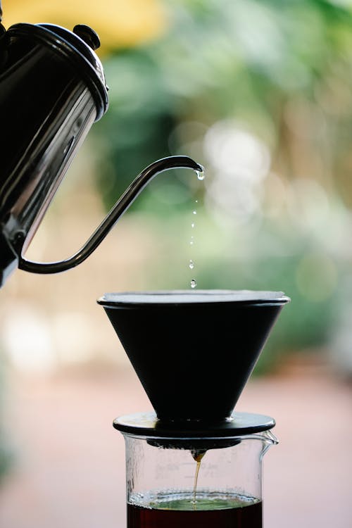 Anonymous person brewing pour over coffee while filling water from kettle into glass coffeemaker in green garden