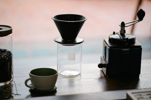 From above of manual coffee grinder with pour over coffeemaker placed in wooden table with ceramic cup and glass jug with roasted beans in morning