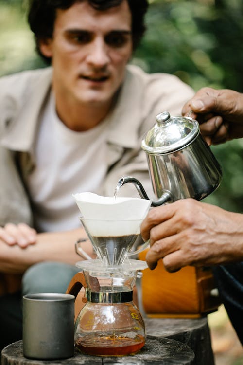 Curious male observing anonymous man pouring hot water from kettle into dripper while making pour over coffee on terrace of cafe on blurred background