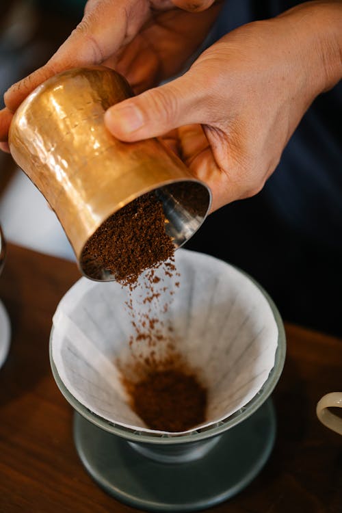 From above of crop anonymous male pouring ground coffee into dripper with filter while making pour over coffee in cafe on blurred background