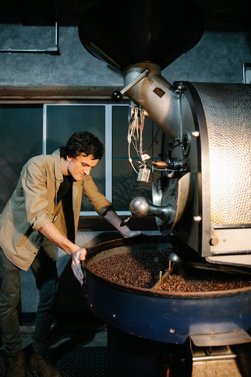 Side view of pensive male standing near modern professional coffee roasting machine with heap of coffee beans in rotating drum