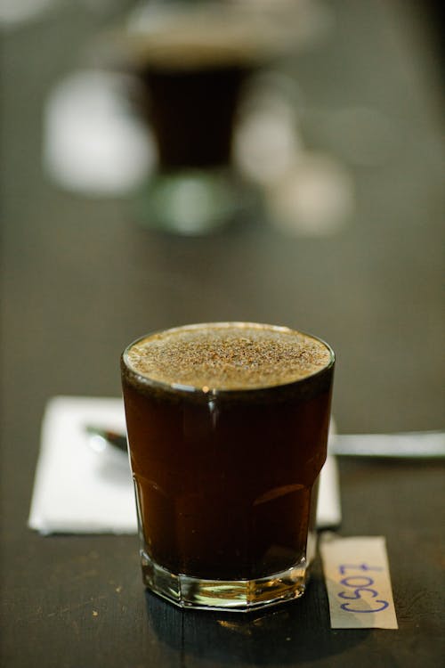 Glass cup of freshly brewed coffee with froth placed on table with spoon and piece of paper with numbers on blurred background during cupping