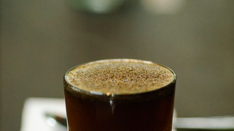 Glass cup of freshly brewed coffee with froth placed on table with spoon and piece of paper with numbers on blurred background during cupping