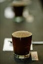 Glass cup of freshly brewed coffee with froth placed on table with spoon and piece of paper with numbers on blurred background during cupping