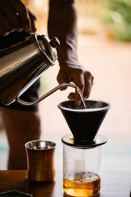 Crop barista pouring hot water from kettle into dripper while preparing alternative pour over coffee