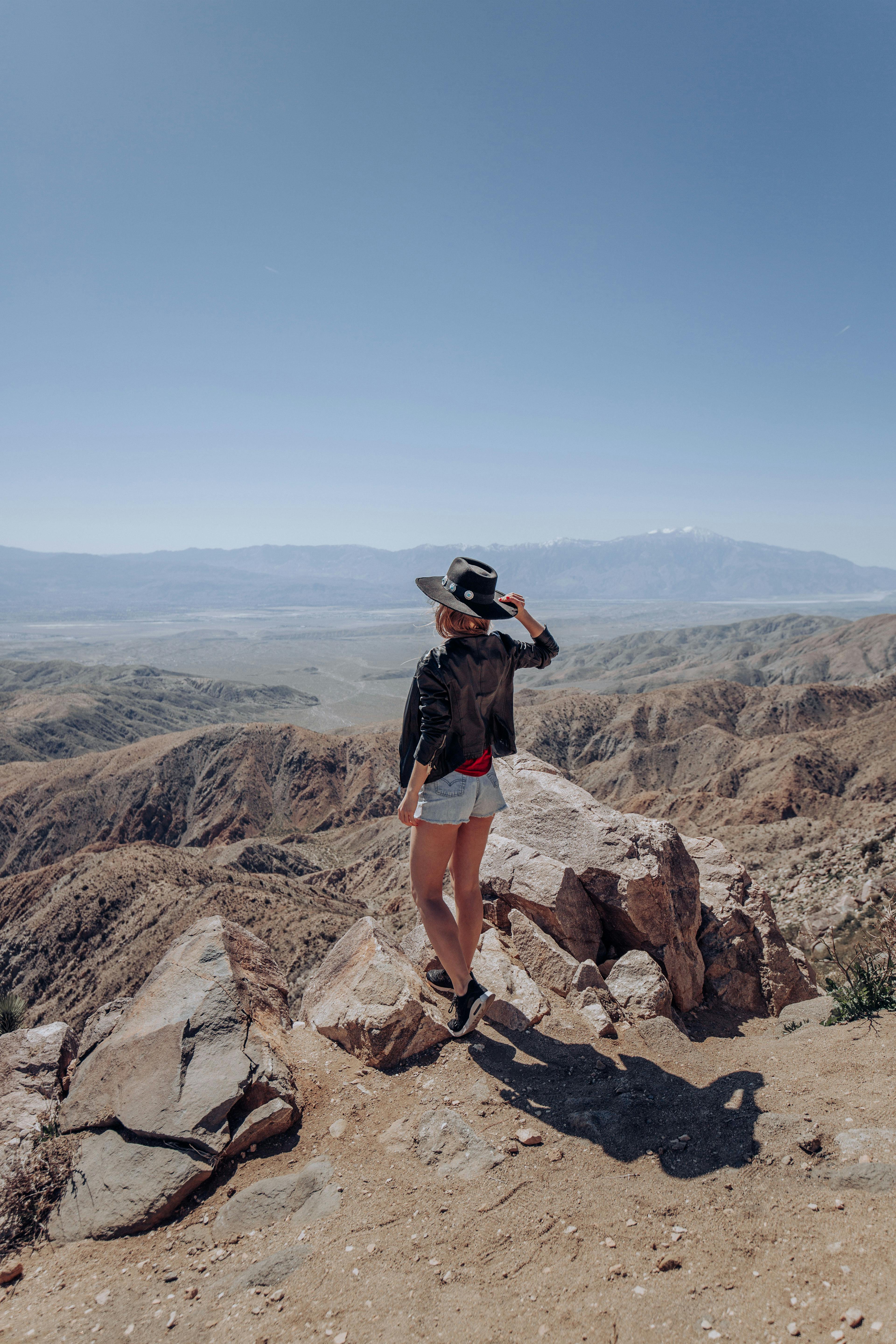 A woman in a black hat and jacket stands on a rocky cliff, overlooking a vast desert landscape.