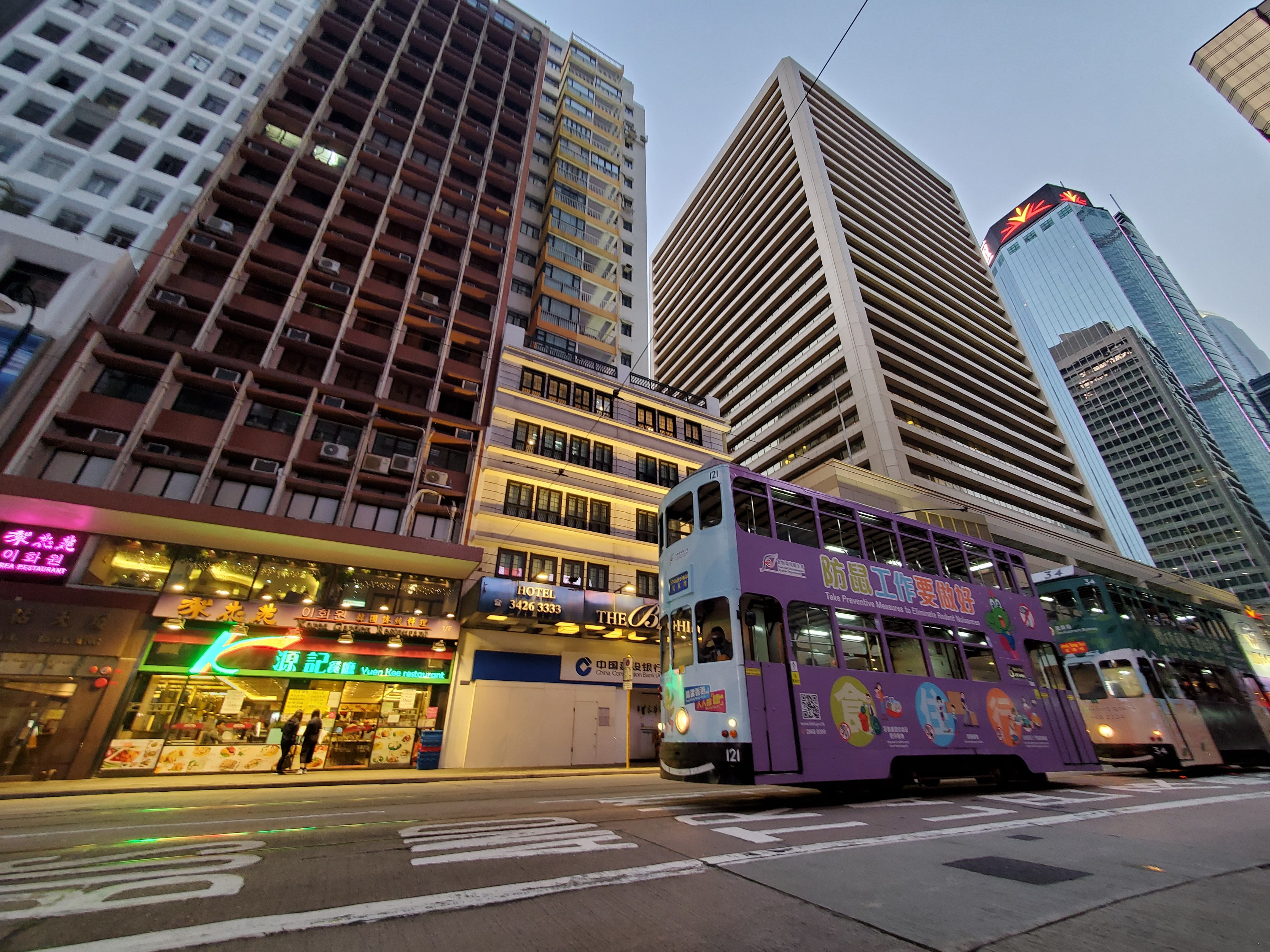 A colorful tram travels down a city street lined with high-rise buildings illuminated at dusk.