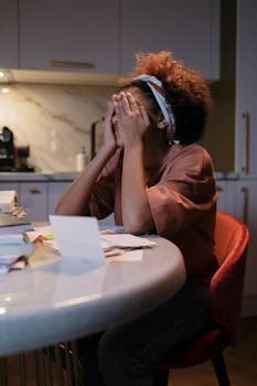 A woman with curly hair sits at a kitchen table looking tired, with hands on face.