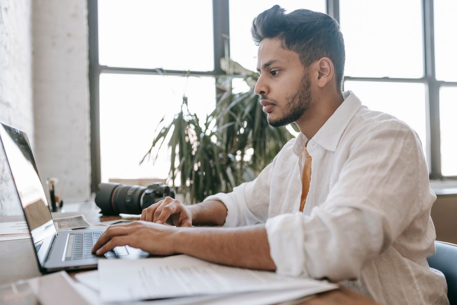 Side view attentive young ethnic male photographer wearing white shirt editing photos on modern netbook while sitting at desk with professional photo camera
